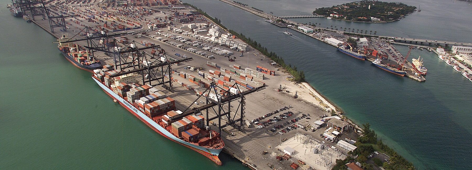 Aerial view of a United States container port, with a container ship berthed alongside gantry cranes and a yard of stacked containers